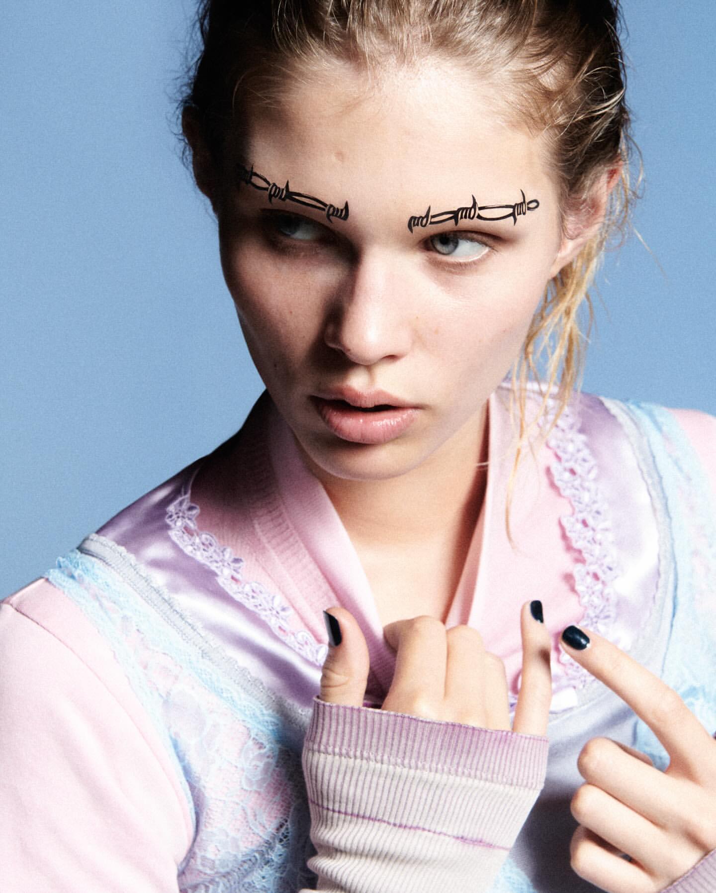 Close-up of a young woman with barbed wire eyebrow art, wearing a pink and blue satin top with striped arm warmers, against a blue backdrop.