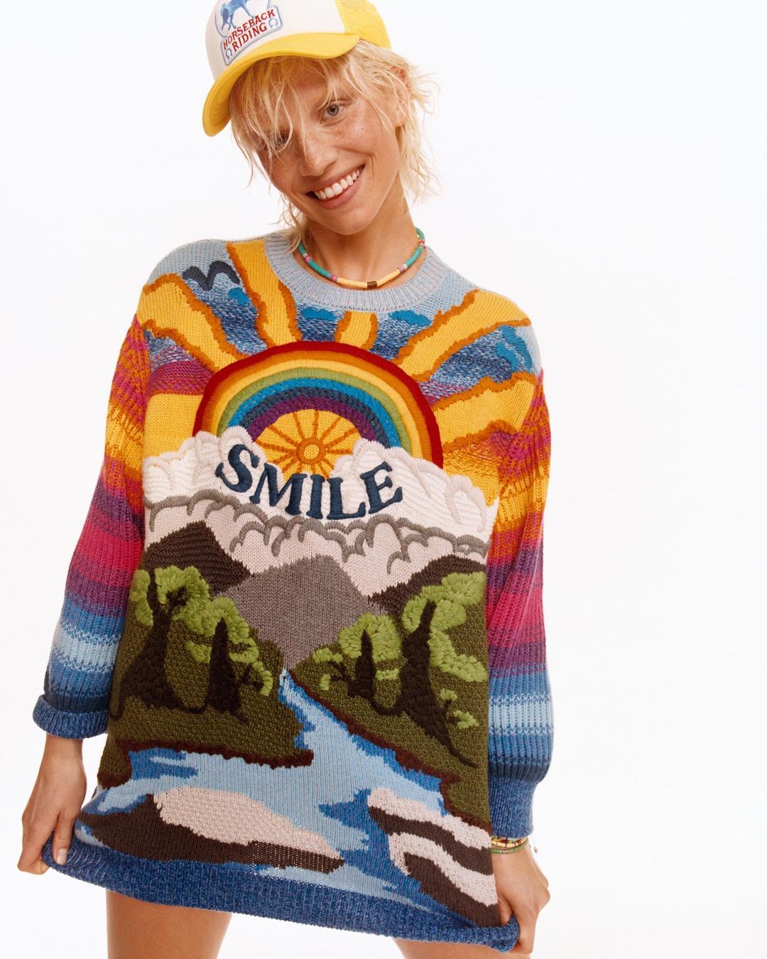 Smiling woman in a yellow hat and colorful sweater featuring landscape art, rainbow, and SMILE typography against a white backdrop.