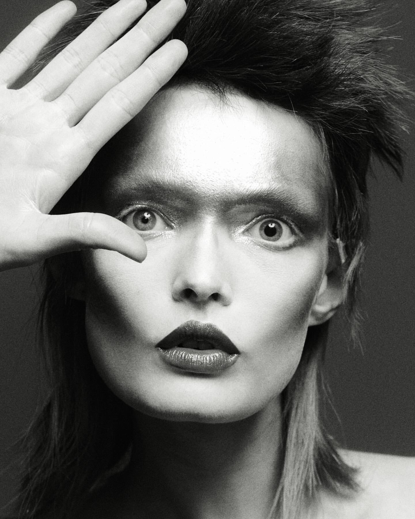 Dramatic black and white portrait of a person with theatrical makeup and styled hair, their hand partially covering their face in studio lighting.