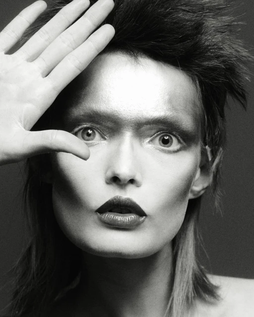 Dramatic black and white portrait of a person with theatrical makeup and styled hair, their hand partially covering their face in studio lighting.
