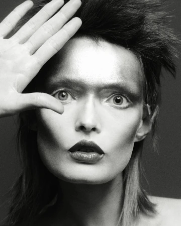 Dramatic black and white portrait of a person with theatrical makeup and styled hair, their hand partially covering their face in studio lighting.