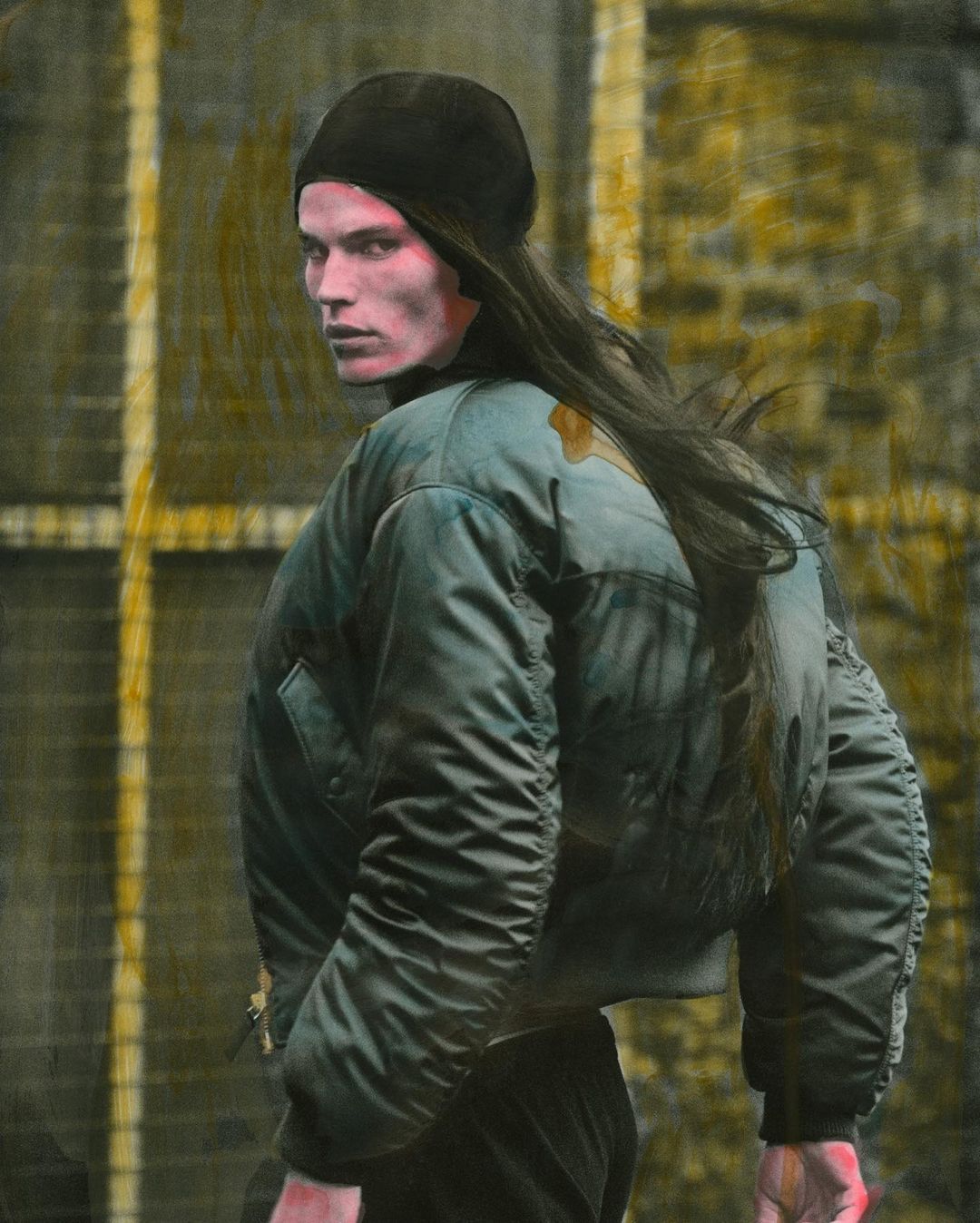 A man with long hair wearing a black beanie and olive green jacket looks over his shoulder in an artistic portrait against a yellow background.