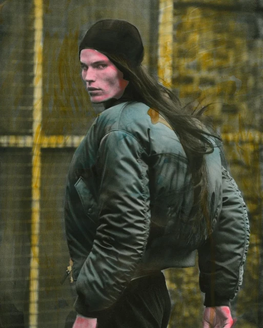 A man with long hair wearing a black beanie and olive green jacket looks over his shoulder in an artistic portrait against a yellow background.
