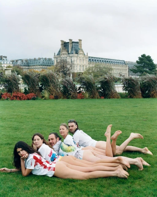Five people, mostly nude, lie on green grass near a building. The group wears white t-shirts. The setting appears to be a park.