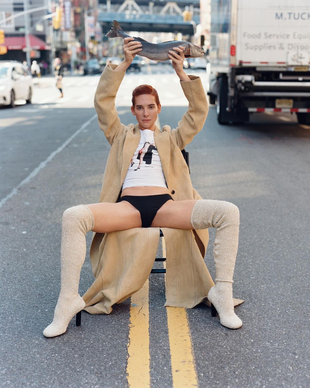 A slender adult with auburn hair sits on a chair in the middle of a street holding a fish, while wearing a tan coat and thigh-high socks.