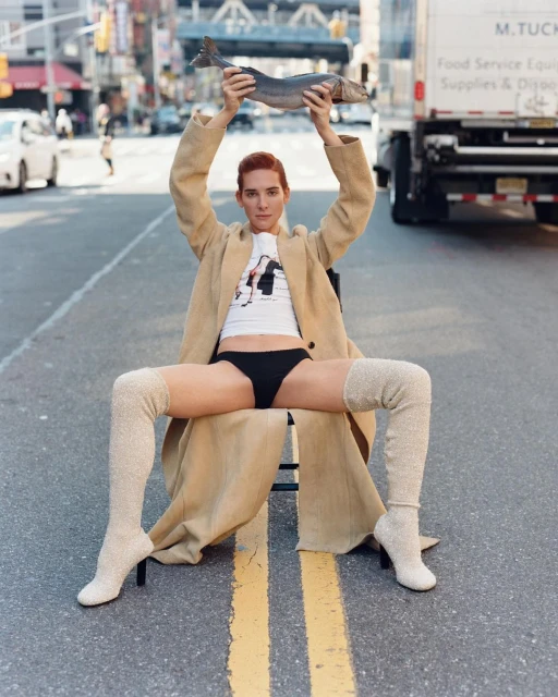 A slender adult with auburn hair sits on a chair in the middle of a street holding a fish, while wearing a tan coat and thigh-high socks.