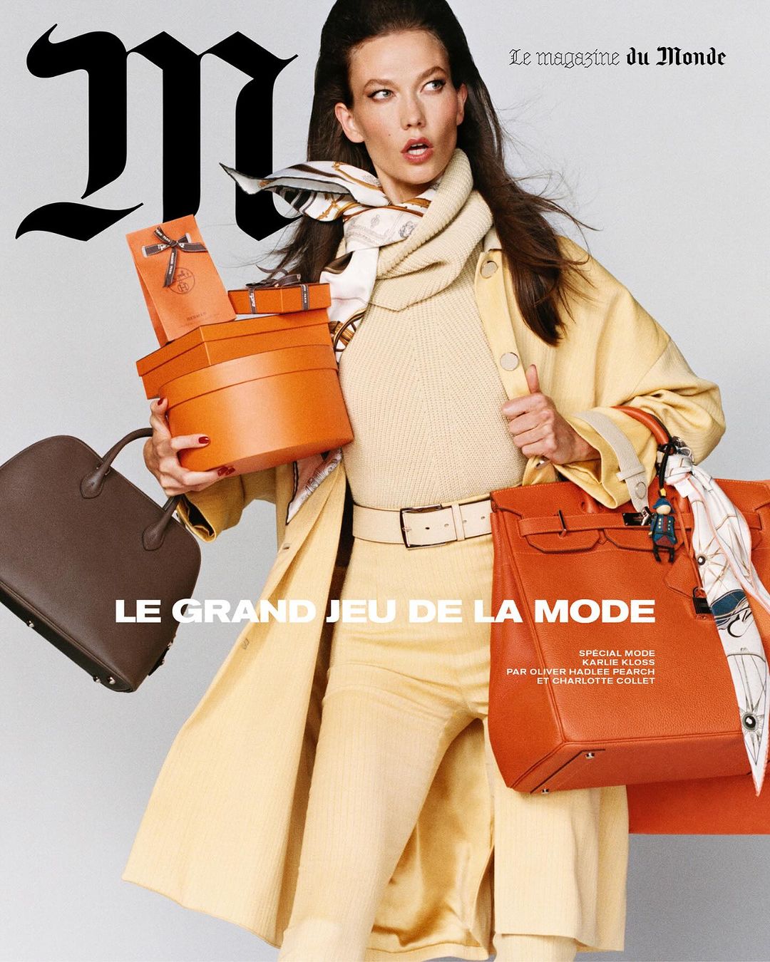 Model Karlie Kloss carrying multiple orange gift boxes and handbags wearing a yellow coat against a grey background