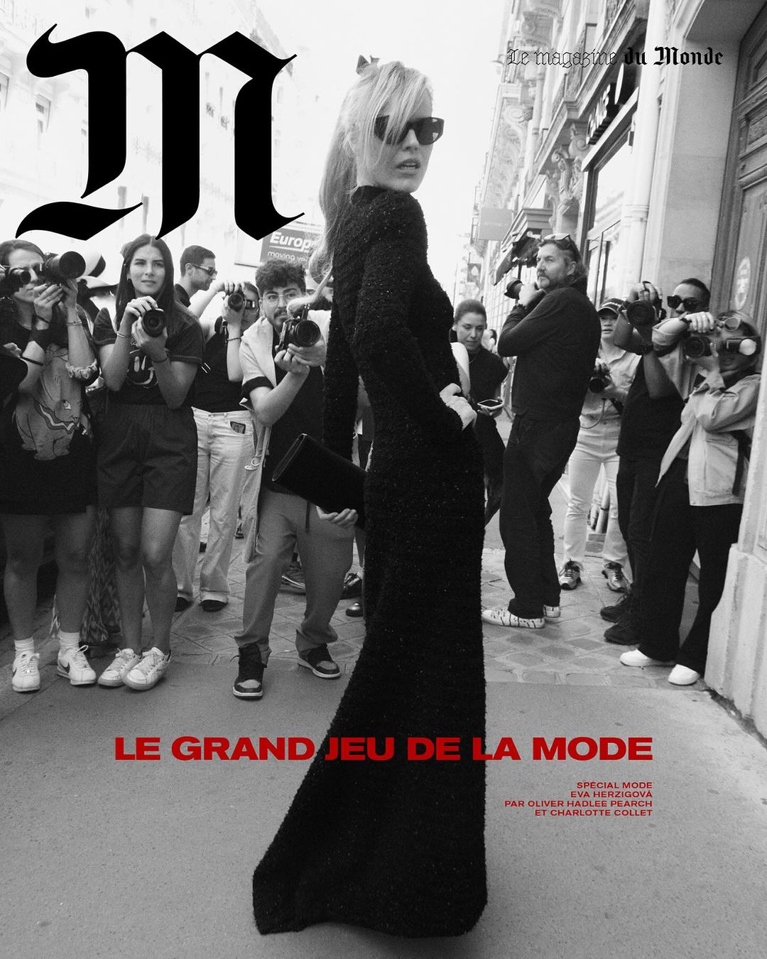 Black and white magazine cover shot outdoors featuring a woman in a black dress, surrounded by photographers and a large letter 'M' logo.