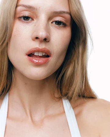 Close up portrait of a young woman with blonde hair and glossy lips wearing a white top against a clean white background