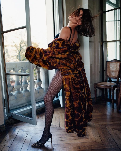 Fashionable woman in robe and heels posing by window with natural light, showing fishnet stockings and elegant details.