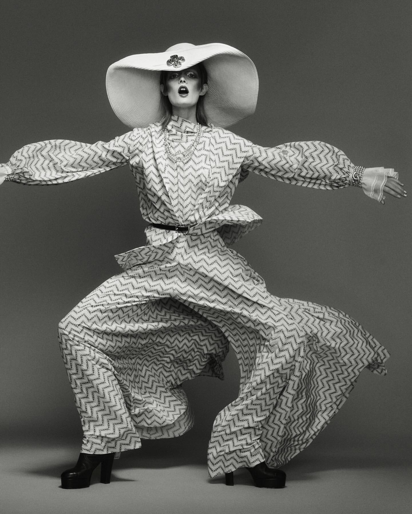 A striking black and white fashion shot of a model in a patterned outfit, wide hat, and platform boots against a gray studio backdrop.