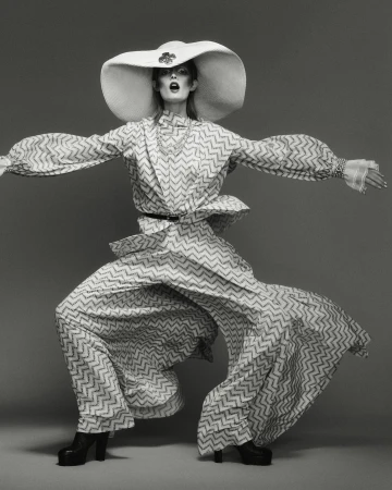 A striking black and white fashion shot of a model in a patterned outfit, wide hat, and platform boots against a gray studio backdrop.