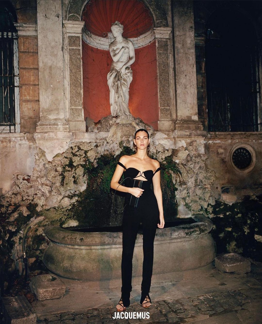 Woman in black dress poses near stone fountain and statue at night. Fashion shot with classic European architecture.
