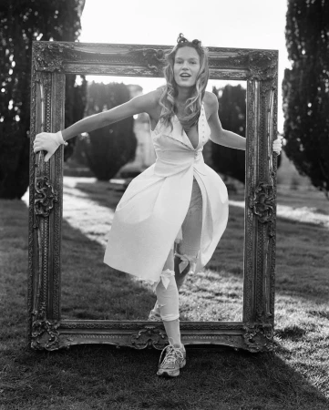A person poses in a dress and sneakers, holding a large ornate empty picture frame in a grassy yard with trees under bright daylight.