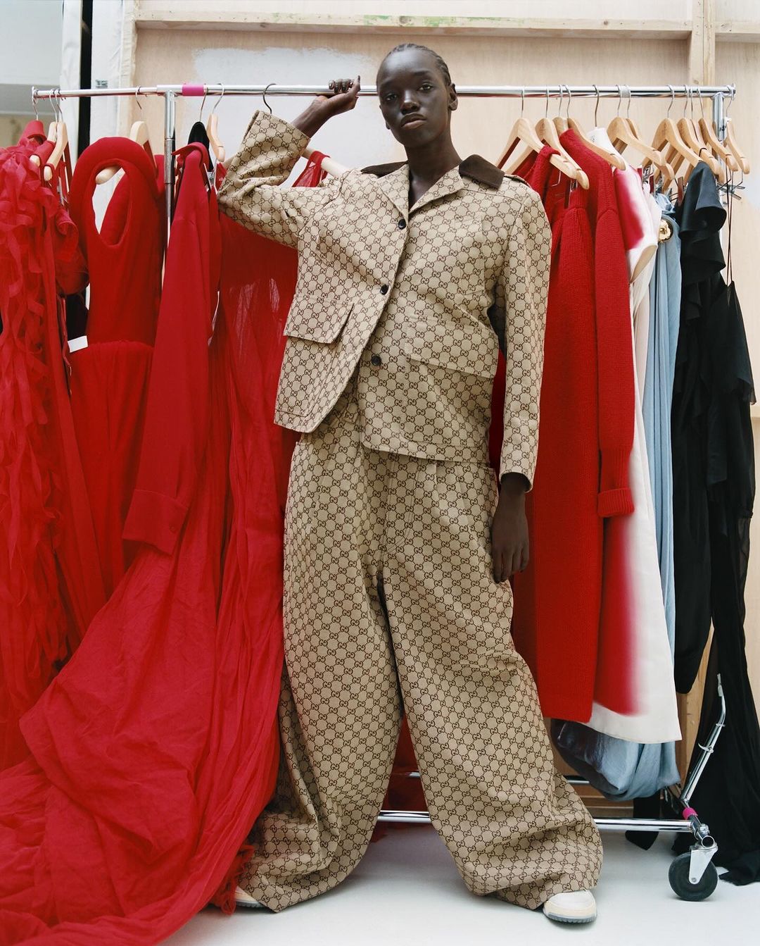 Woman in patterned suit poses near rack of red dresses in a fashion shot, indoor lighting, eye-level angle, studio or backstage environment.