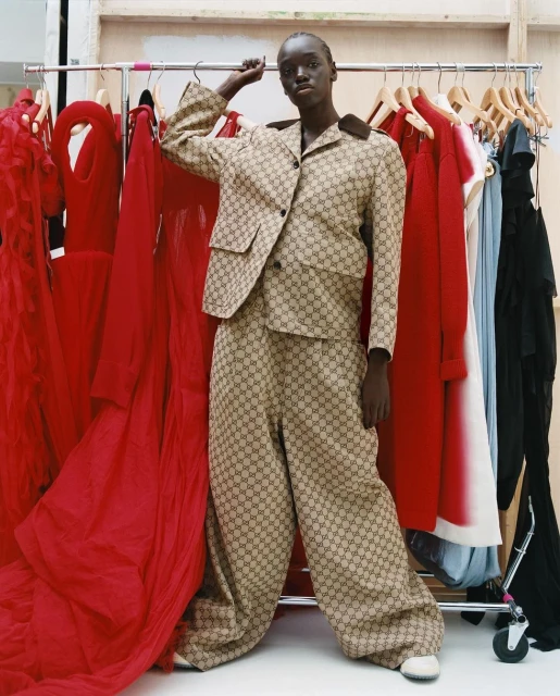 Woman in patterned suit poses near rack of red dresses in a fashion shot, indoor lighting, eye-level angle, studio or backstage environment.