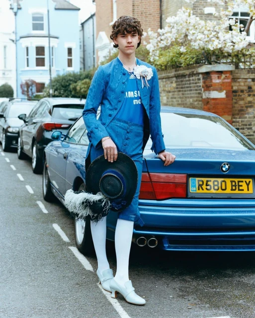 Young adult stands by a blue BMW in an ornate blue costume, holding a feathered hat on a tree-lined urban street on a sunny day.