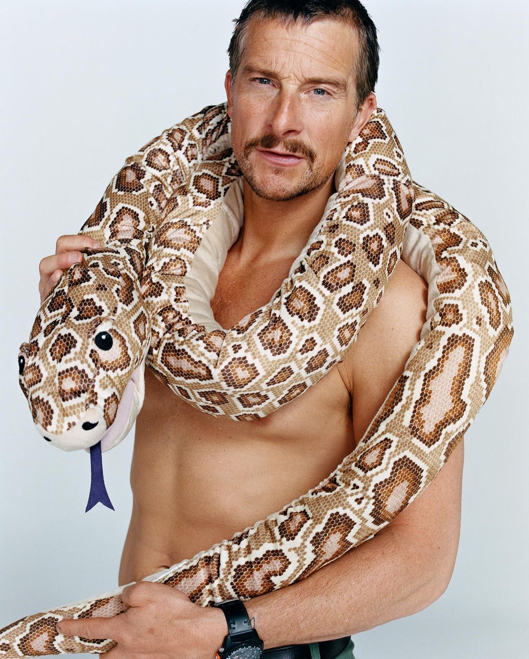 Adult male posing indoors with a toy snake wrapped around his shoulders, set against a clean white backdrop in a portrait shot.