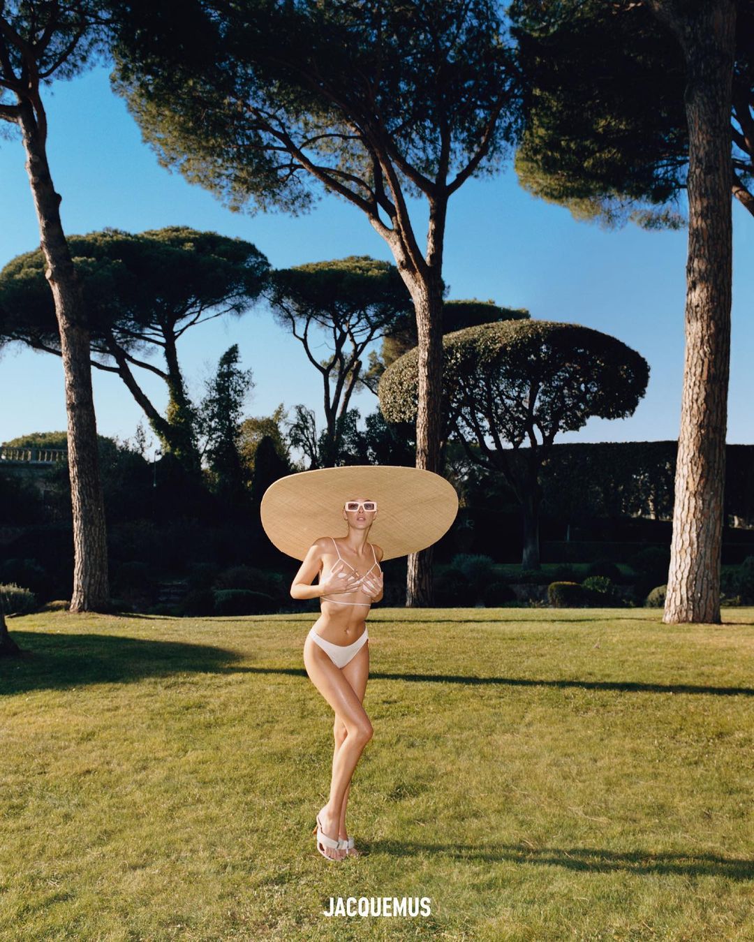 A woman in a bikini and large hat stands on a lawn surrounded by trees, bathed in sunlight, promoting fashion with a luxury feel.