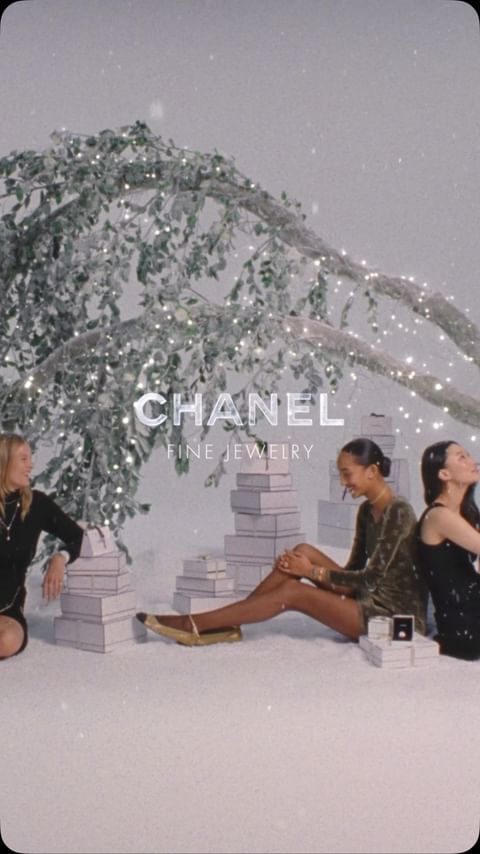 Three women sit among stacked gift boxes near a tree with fairy lights in a holiday-themed fashion and jewelry advertisement.