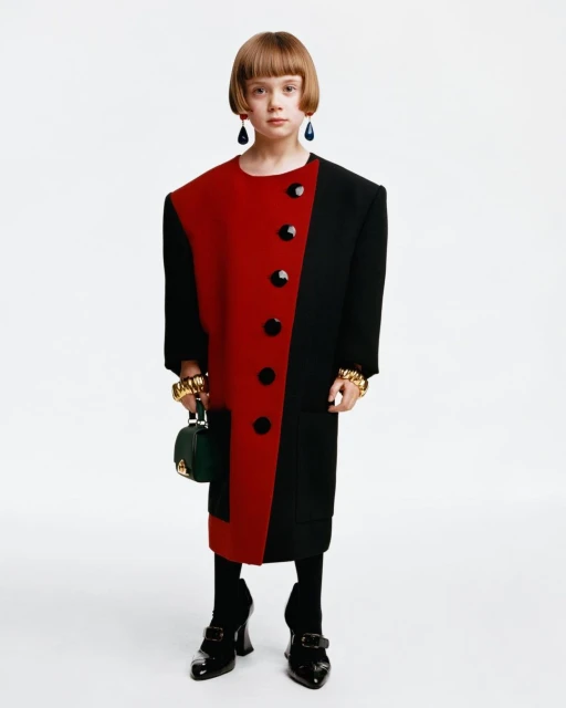 Child model in a color-blocked red and black oversized coat dress with black tights, patent leather shoes, green handbag and gold jewelry.