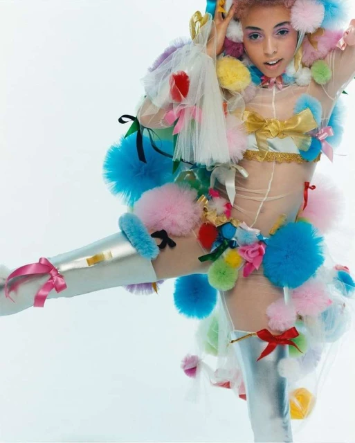Woman in avant-garde fashion poses in a sheer bodysuit with colorful pom poms, ribbons, and metallic thigh-high boots against a white backdrop.