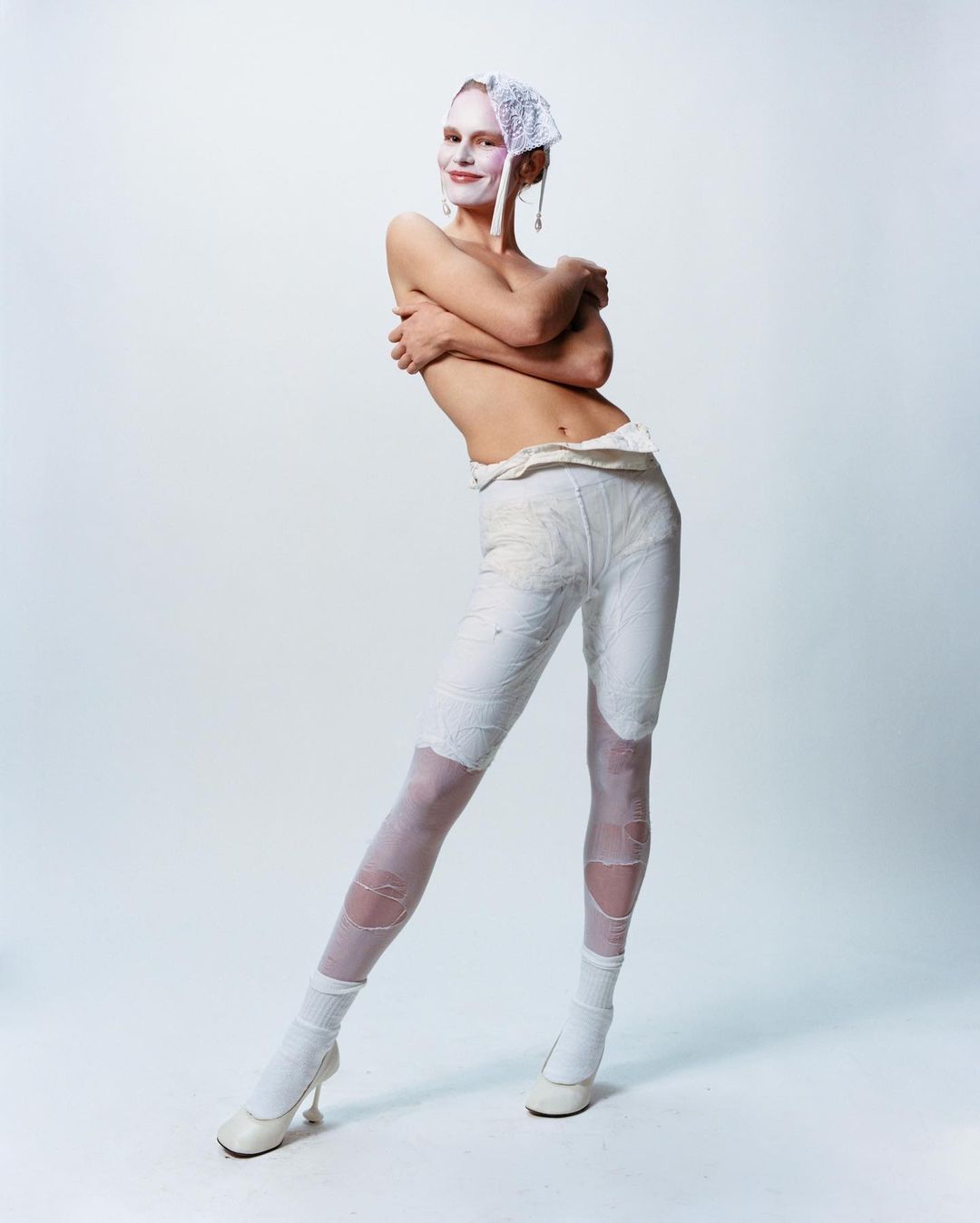 Full body portrait of an adult female in a white outfit and cap with white makeup, posing against a white studio background.