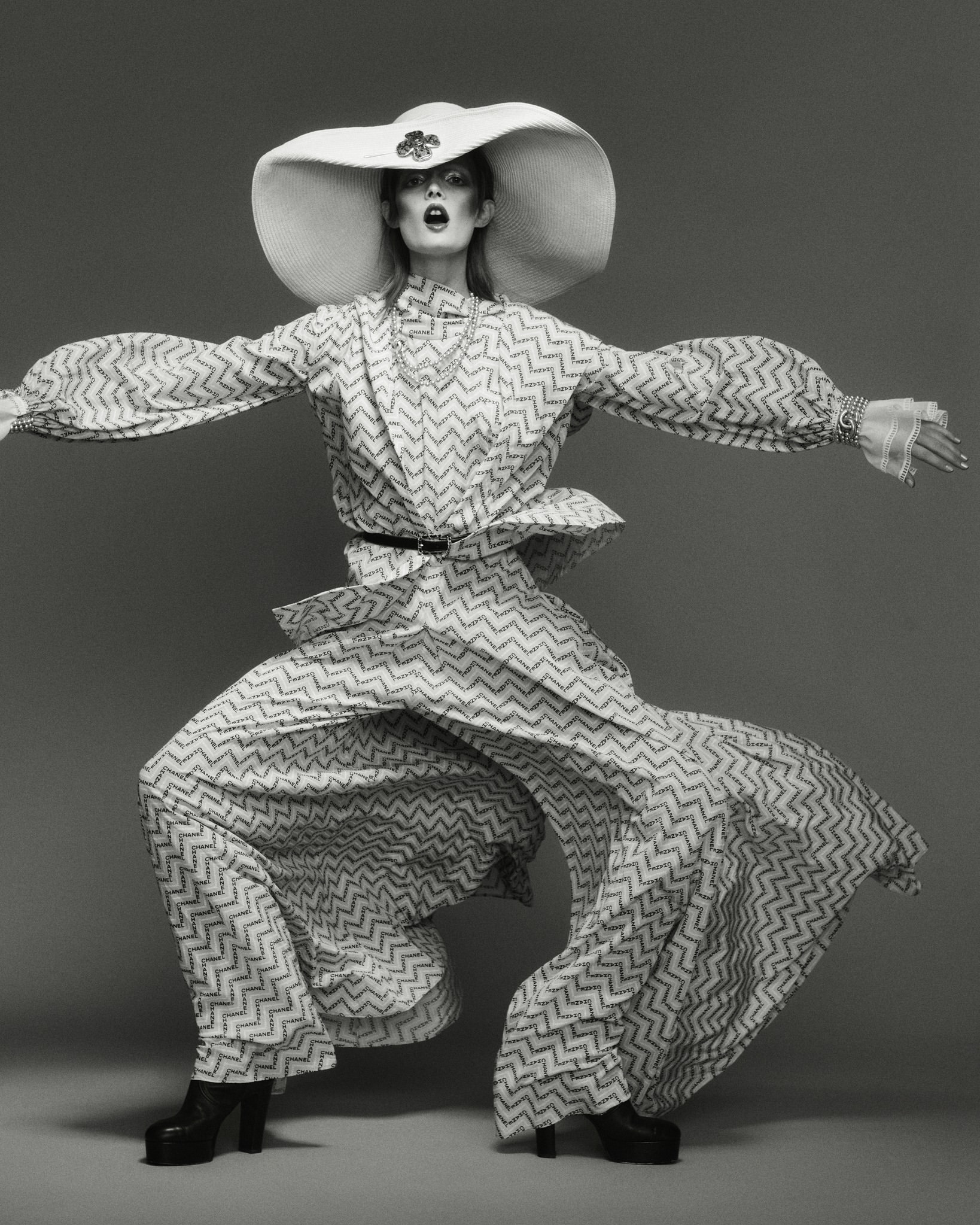 Woman in oversized patterned suit and hat poses in studio. Fashion shot in grey tones showcasing retro-inspired design and silhouette.