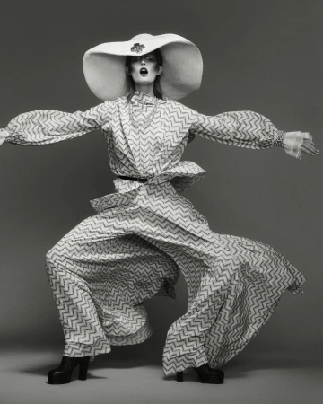 Woman in oversized patterned suit and hat poses in studio. Fashion shot in grey tones showcasing retro-inspired design and silhouette.