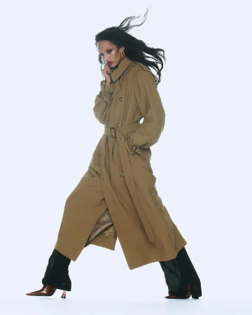 Model in long tan trench coat walking against a bright white background with dark hair blowing