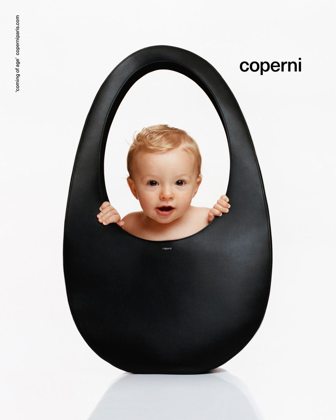A baby with blonde hair peeks out of a large black Coperni bag against a white background in a studio shot for a fashion advertisement.
