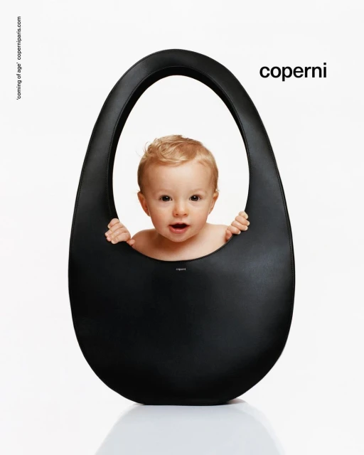 A baby with blonde hair peeks out of a large black Coperni bag against a white background in a studio shot for a fashion advertisement.