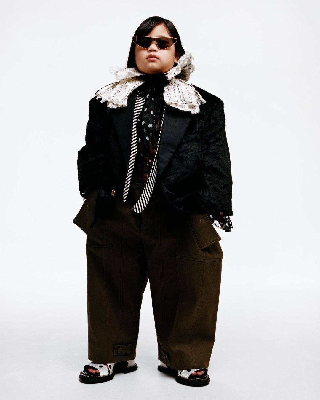 Fashion-forward individual with sunglasses poses in oversized blazer, ties, and cargo pants against a clean white backdrop.