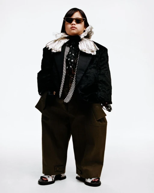 Fashion-forward individual with sunglasses poses in oversized blazer, ties, and cargo pants against a clean white backdrop.