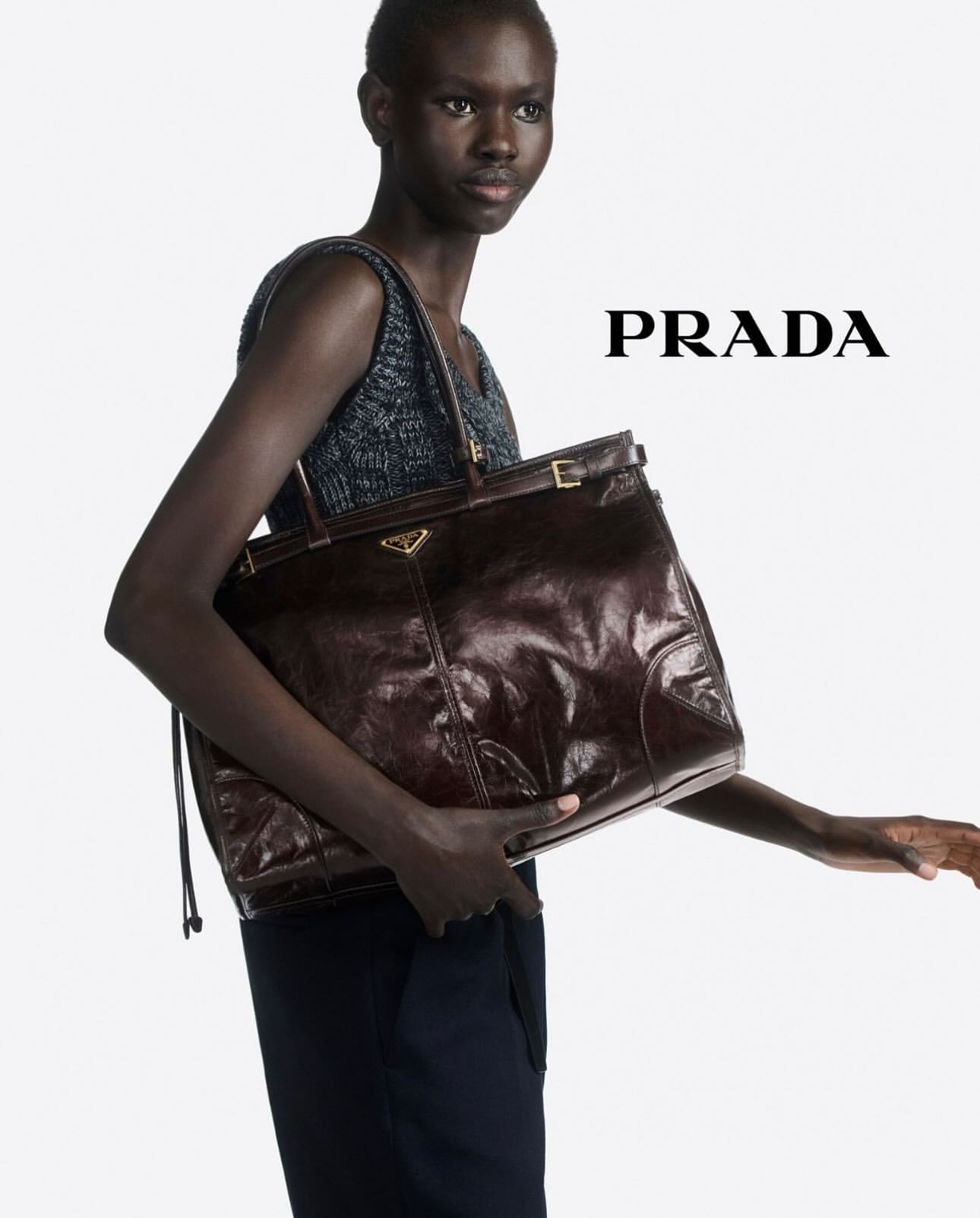 Model holding a large dark brown leather tote bag against a bright white background with Prada text