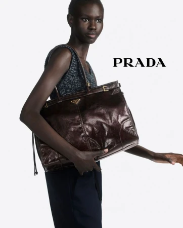 Model holding a large dark brown leather tote bag against a bright white background with Prada text