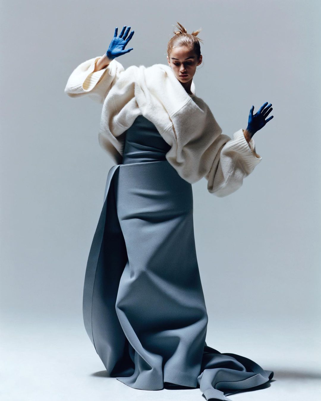 Fashion model in a voluminous white sweater and long blue skirt poses with hands raised against a solid grey background.