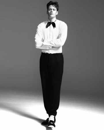 Monochrome portrait of a man standing with arms crossed wearing a white shirt, black bow tie, and dark trousers.