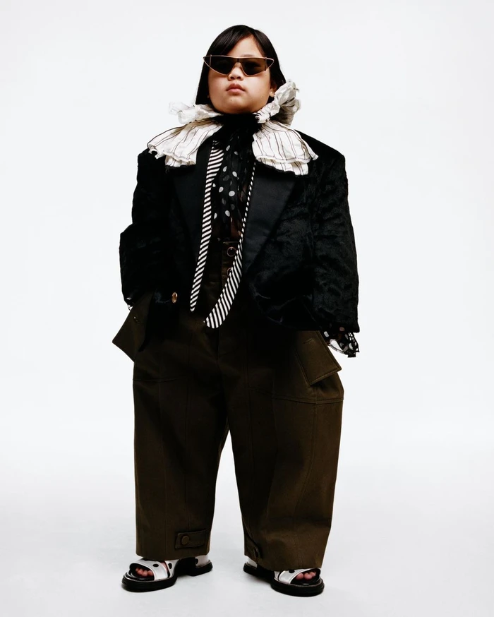 Child wearing oversized black blazer, ruffled collar, sunglasses and wide trousers against a white backdrop