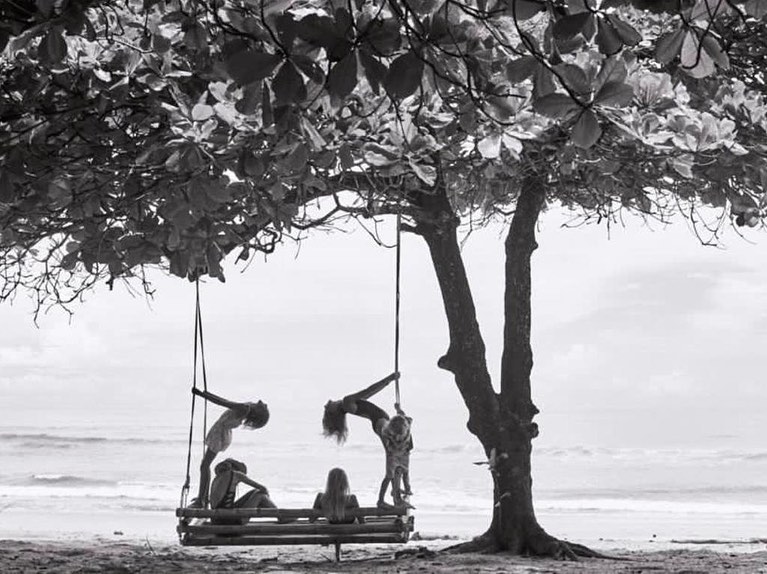 Black and white photo of five young women on a wooden swing hanging from a mature tree on a beach, with the ocean in the background.