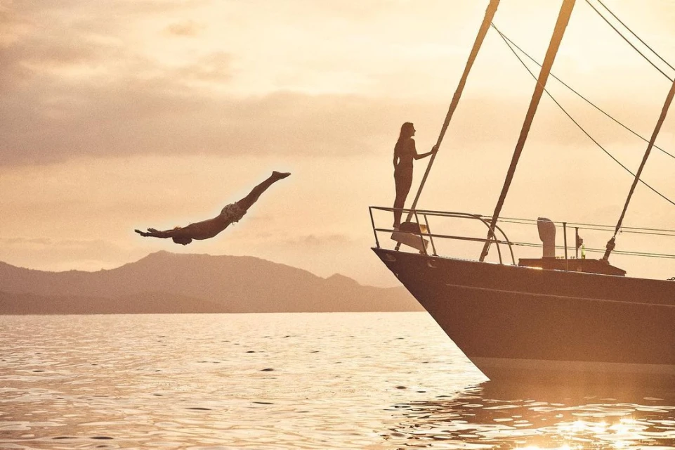 Man dives from a sailboat on the water at sunset with a woman standing on the bow, silhouetted mountains in the background.