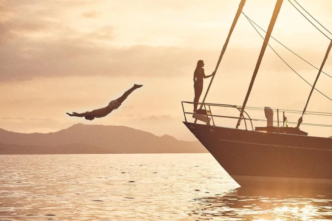 Man dives from a sailboat on the water at sunset with a woman standing on the bow, silhouetted mountains in the background.