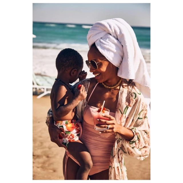 A woman in swimwear and a towel on her head smiles while holding a child on a beach with the ocean in the background.