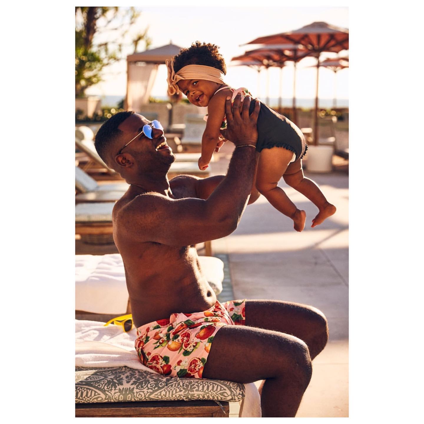 Father and child in swimwear enjoy a sunny day by the pool, with the father holding the child in the air and smiling.