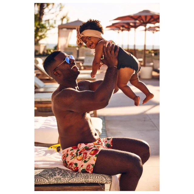 Father and child in swimwear enjoy a sunny day by the pool, with the father holding the child in the air and smiling.