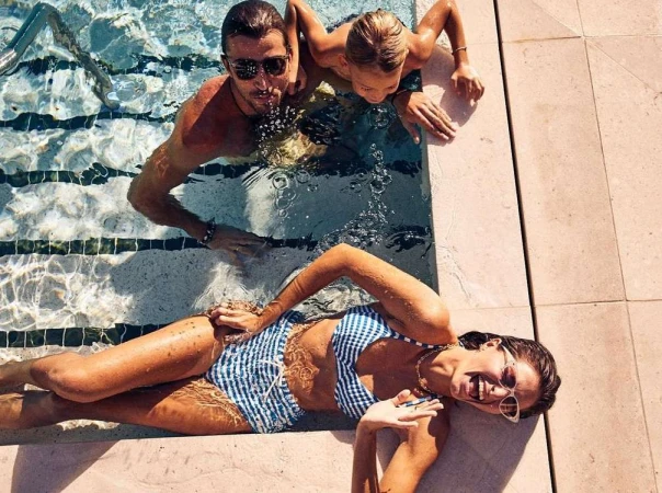 Woman in a blue and white swimsuit relaxes on the edge of a pool with a man and child visible in the water, shot from overhead.