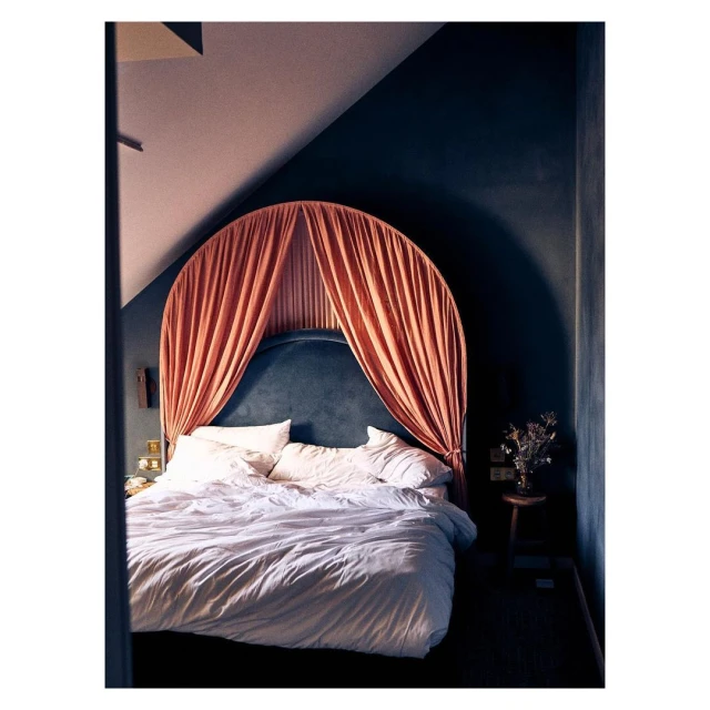 Canopy bed with white bedding in a dark blue room with bedside table and wall lighting provides a cozy, intimate interior design.