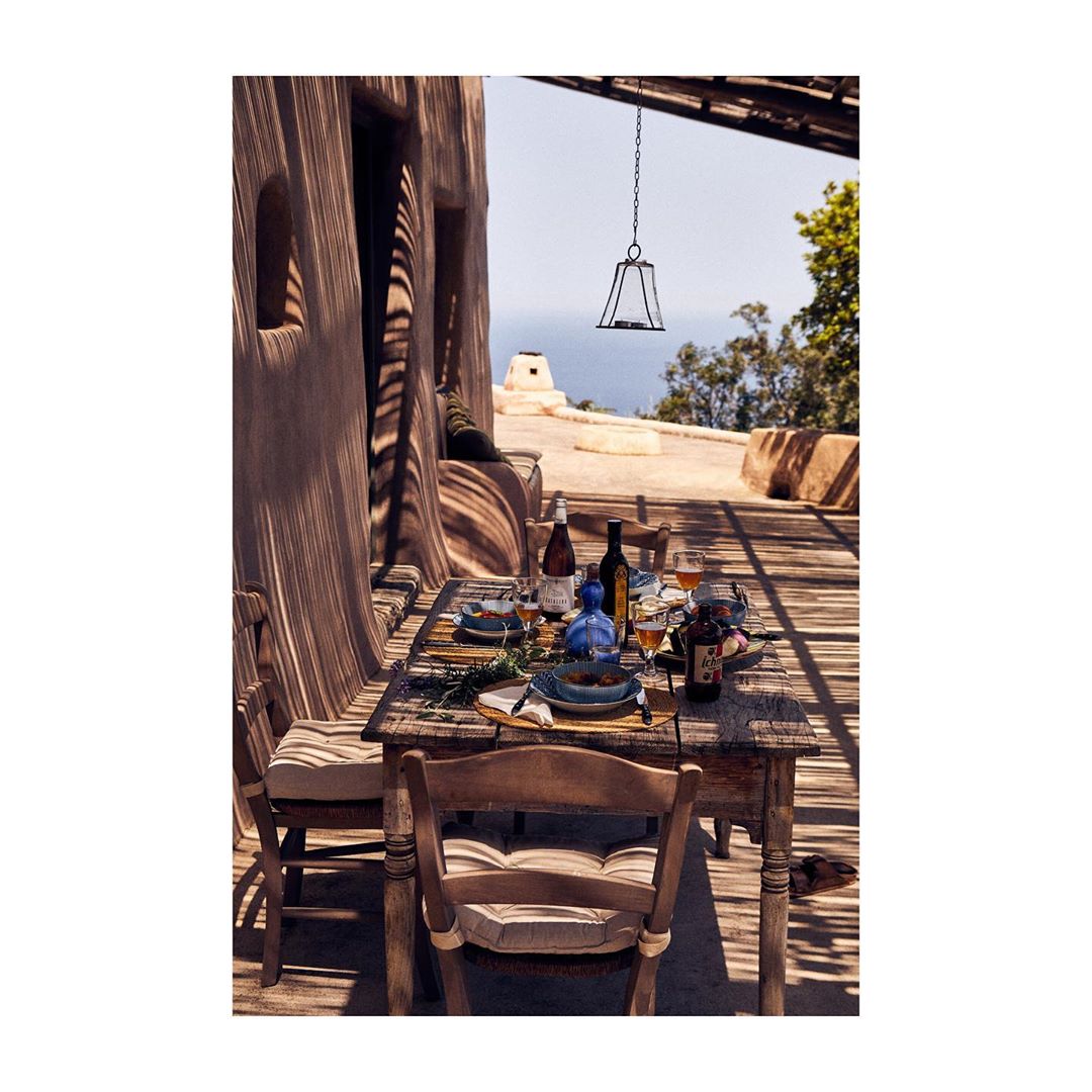 Outdoor dining table on a patio with bottles, glasses, plates, and silverware set for a meal, with chairs and shadows visible.