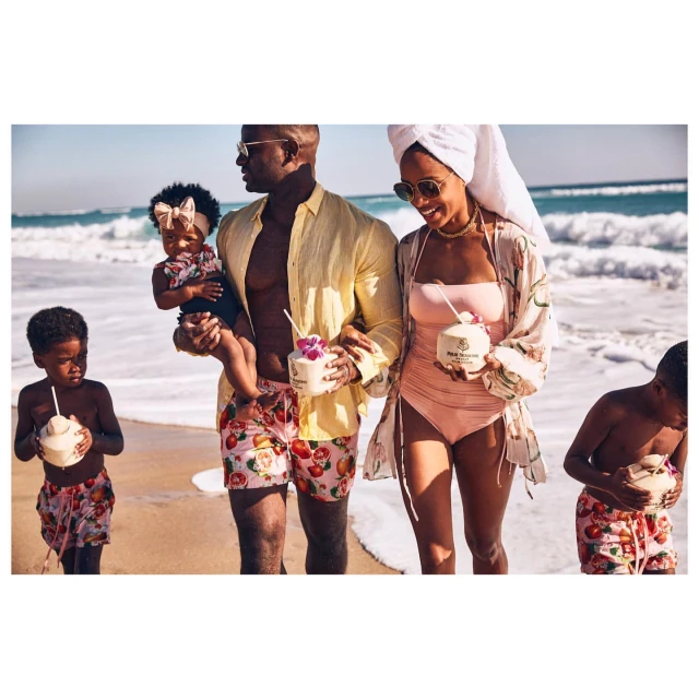 Family of four on a sandy beach with waves. Two adults and two children enjoy coconut drinks on a sunny day by the ocean.