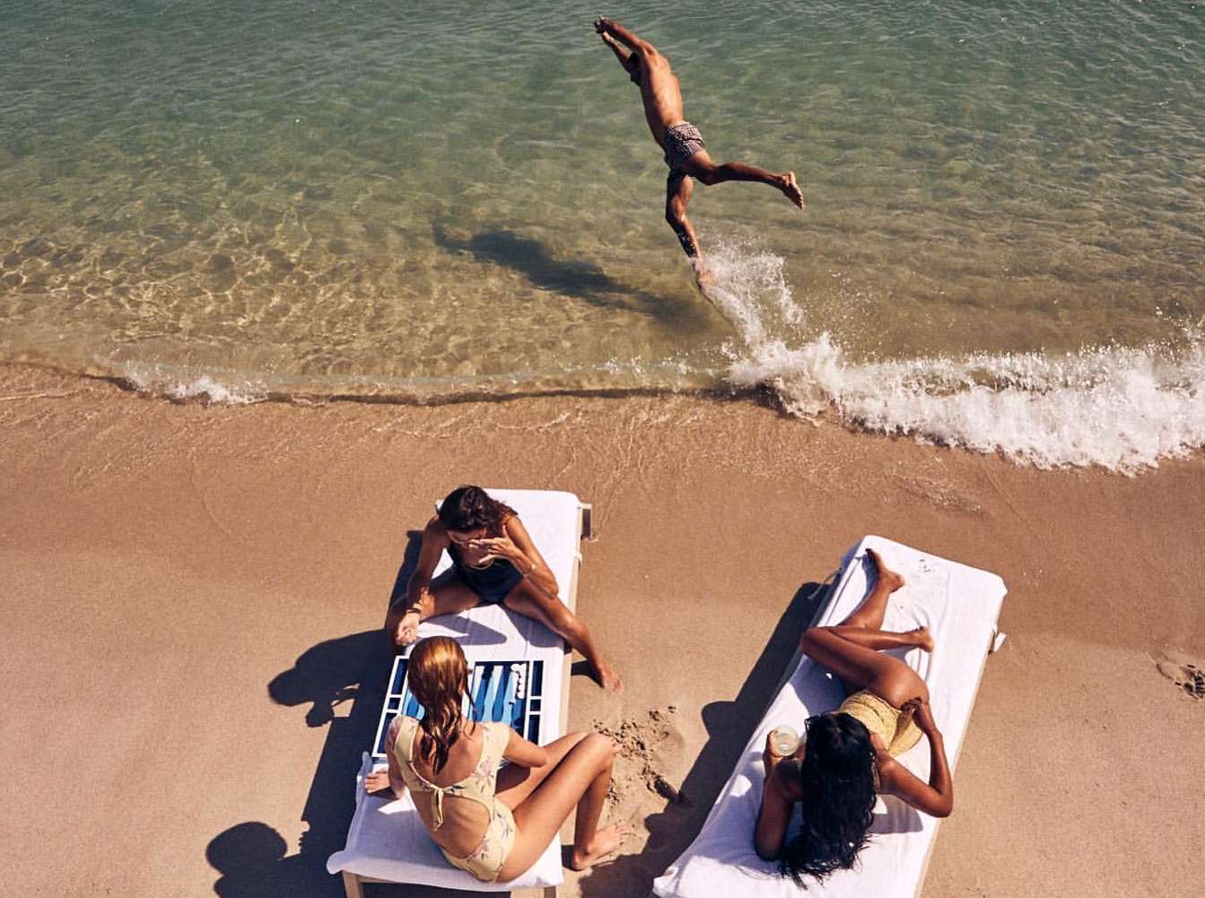 Beach scene with three women on lounge chairs and one man jumping over the ocean waves on a sunny day, capturing a summer vibe.